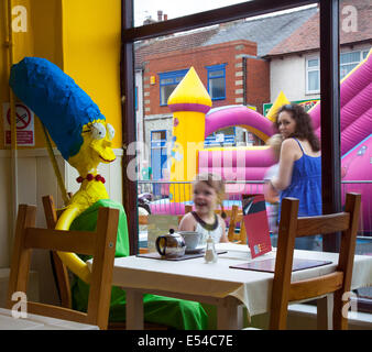 Marge Simpson Fancy-Kleid Fleetwood, Lancashire, 20. Juli 2014. Freya & Heidi Pollit, die durch ein Café-Fenster bei Marjorie „Marge“ Simpson schauen, tragen einen erwachsenen Blue Wig, Yellow 2. Skin Supersuita fiktionaler Charakter in der amerikanischen animierten Sitcom, beim Fleetwood Festival of Transport. UK Stockfoto