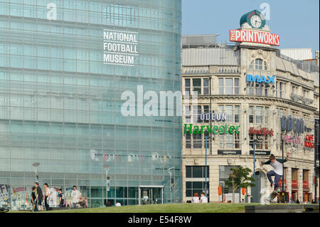 Ein junger Mann versucht einen Skateboard Trick vor dem National Football Museum (ehemals Urbis) und Printworks in Manchester. Stockfoto