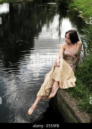 Junge Frau im Sommerkleid saß am Ufer des Flusses mit den Zehen im Wasser Stockfoto