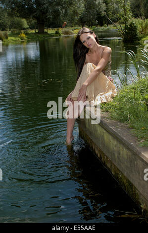 Junge Frau im Sommerkleid saß am Ufer des Flusses mit den Zehen im Wasser Stockfoto