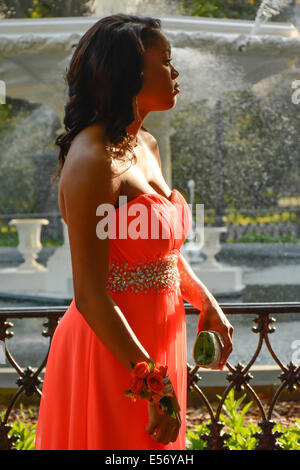 Eine schöne junge afroamerikanische Frau im Profil trägt Orange Ballkleid mit einem Handgelenk Corsage vor dem Brunnen in Forsyth Park, Savannah, GA, USA Stockfoto