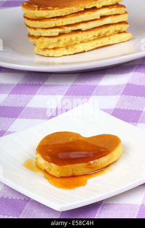 Mini Herzform Pfannkuchen mit Sirup auf einem weißen Teller. Im Hintergrund ein Stapel von Pfannkuchen. Ein perfektes Frühstück zum Valentinstag Stockfoto