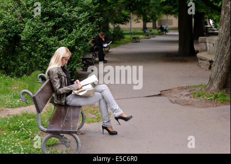 Eine junge modische schlanke, blonde Frau sitzt auf einer Bank und liest ein Buch in einem Park, Prag, Tschechische Republik. Stockfoto
