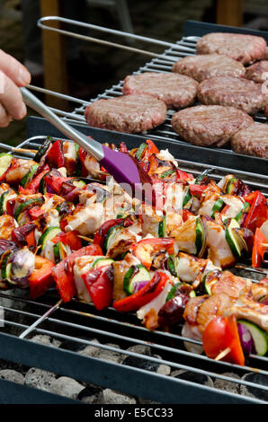 Holzkohlegrill BBQ mit Hähnchenspieße und Beef Burger von einem Mann mit einer Bürste aus Kunststoff Küche begossen wird geladen Stockfoto