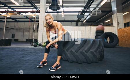 Schöne und starke junge Frau sitzt auf einem großen Reifen nach ihrer Übung. Frau Pause nach Crossfit Training im Fitness-Studio. Stockfoto