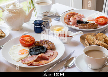 Traditionelles englisches Frühstückstisch ausgebreitet durch Fenster Innenraum mit Tee Toast Speck, Würstchen und Eiern auf Tischdecke Stockfoto