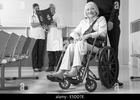 Reife Frau im Rollstuhl mit ihrem Ehemann im Krankenhaus. Ärzte analysieren Scan im Hintergrund. Stockfoto