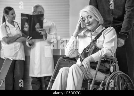 Reife Frau im Rollstuhl im Krankenhaus sorgen mit ihrem Ehemann. Ärzte analysieren Scan im Hintergrund. Stockfoto