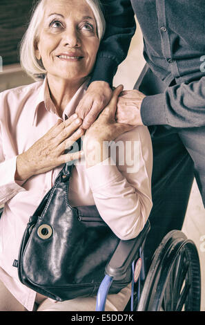 Reife Frau im Rollstuhl lächelnd zu ihrem Mann nimmt seine Hand. Stockfoto