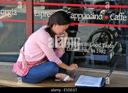 asiatische Frau auf Handy sitzt auf Bank außerhalb Café multitasking Stockfoto