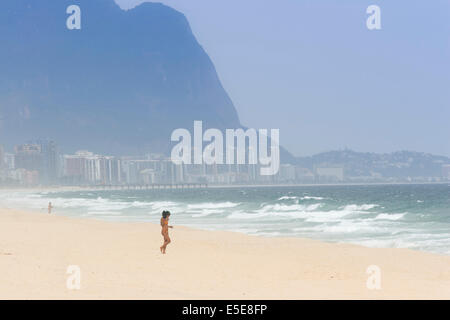 Pontal, Barra da Tijuca, Rio. Eine junge Frau in einem Bikini, die mit dem Berg Pedra da Gavea und der Skyline der Stadt in der Ferne in die Wellen läuft Stockfoto