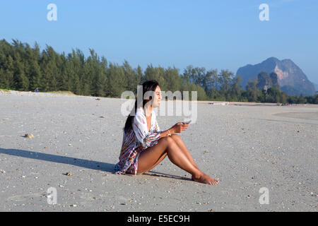 Eine einzelne junge asiatische Frau lächelnd, als sie eine SMS-Nachricht an einem Strand in Thailand sitzen liest Stockfoto
