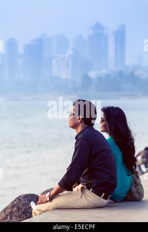 Ein junges Paar sitzt an der Wand neben Marine Drive mit den Türmen der Malabar Hügeln hinter, Mumbai, Indien Stockfoto