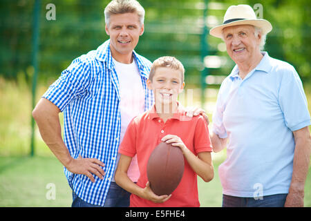 Süsser Boy mit Rugby-Ball und seinem Großvater und Vater Blick auf Kamera im freien Stockfoto