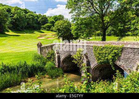 Eine steinerne Fußgängerbrücke über den Fluss von Bach mit einem Golfplatz jenseits. Stockfoto