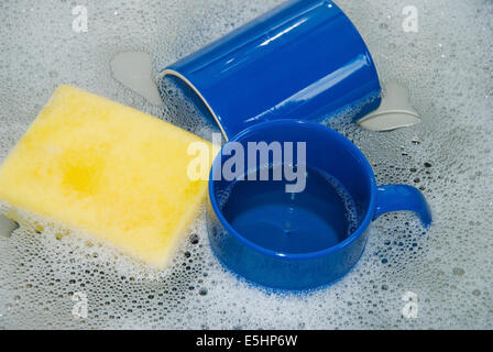 zwei blaue Kaffeetassen mit einem gelben Schwamm in einer Küchenspüle mit Schale Wasser und Schaum, Nahaufnahme Stockfoto