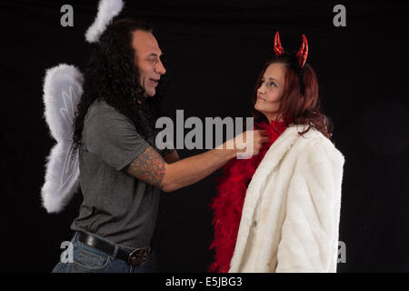 Mann in Engelsflügel und weiße Halos, mit einer Frau in Teufelshörnern und rote Federboa und weißen Pelzmantel Stockfoto