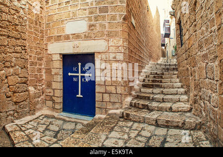 Metalltür mit weißen Kreuz am Eingang zur kleinen Kirche und schmalen gepflasterten Straßen in Jaffa, Israel. Stockfoto