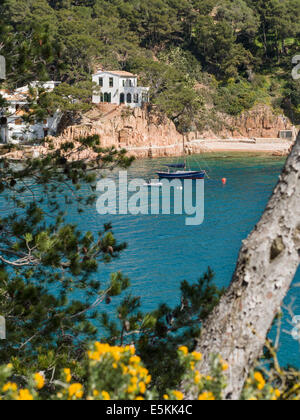 Villa und Segelboot an der Costa Brava. Eine Villa sitzt oben auf einem kleinen Felsen über eine türkisfarbene Bucht mit einem blauen Segelboot Stockfoto