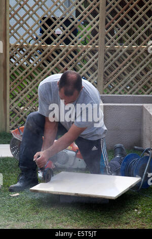 Arbeiter schneiden und legen Terrassenfliesen in Southport, Merseyside, Großbritannien. August 2014. Schneiden von Fliesen mit einem Angle Grinder als Flower Show Vorbereitungen auf den Victoria Park showgrounds, für die größte unabhängige Blumenschau Großbritanniens. Es wurde ursprünglich im Jahr 1924 von der Gemeinde gegründet, aber seit 1986 wird es von Southport Flower Show Unternehmen betrieben, die eine eingetragene Wohltätigkeitsorganisation ist. Es findet jährlich für vier Tage Ende August statt und hat eine Teilnahme von über 80.000 Personen. Stockfoto