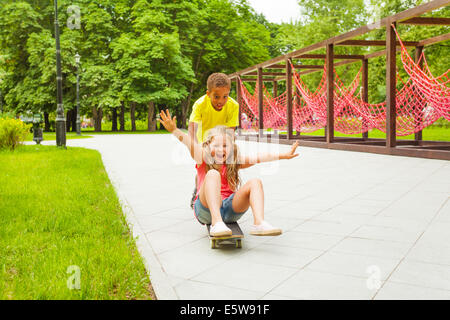 Boy schiebt aufgeregt Mädchen sitzen auf skateboard Stockfoto