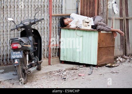 Fotojournalismus Bild der Barefoot burmesischen Mann schlafen auf einer Holzkiste, die in einer Gasse. düstere Bild von echten Menschen und der Realität auf der Straße nach Mandalay Stockfoto