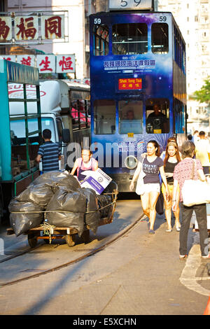 Chinesische Frauen Street Cleaner schiebt einen schweren Warenkorb Müll durch Chun Yeung Street, Vintage Hong Kong Tram im Hintergrund. Stockfoto