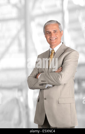 Ein reifer Geschäftsmann mit seinen Armen gefaltet gegen unscharfe moderne Büroeinrichtung. Hochformat. Stockfoto