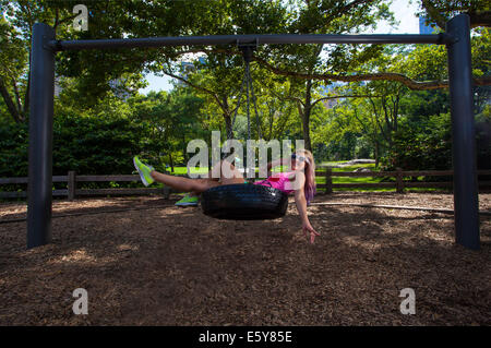 Young blonde sportliche Frau sitzen und Schaukeln auf einer Reifenschaukel im Central Park Stockfoto