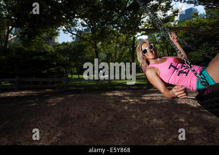 Young blonde sportliche Frau sitzen und Schaukeln auf einer Reifenschaukel im Central Park Stockfoto