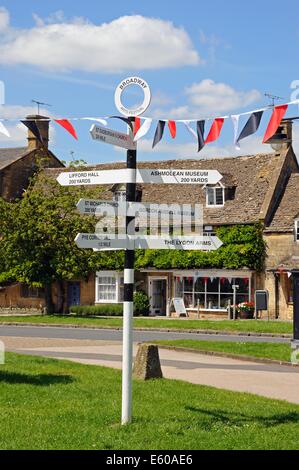 Schwarz / weiß-Wegweiser auf dem Dorfanger entlang der High Street und Wegbeschreibungen zu den touristischen Attraktionen, Broadway, England. Stockfoto