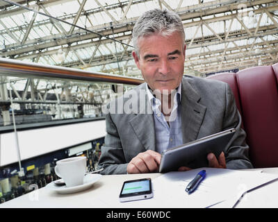 Reifen Geschäftsmann im Cafe in der Tabelle auf der Eisenbahn Halle bei seinem iPad Tablet Computer mit iPhone und Hinweise im Vordergrund suchen sitzt Stockfoto