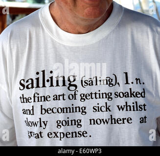 Ein Mann an Bord eines Segelbootes im karibischen Meer trägt ein weißes T-shirt mit Schrift, die eine humorvolle Wörterbuch-ähnliche Definition des Wortes "Segeln" gibt. Stockfoto