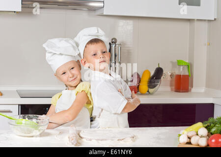 Niedlichen Mädchen und jungen stolz, Chef Uniform zu tragen, während des Backens in der Küche posiert Rücken an Rücken mit verschränkten Armen Stockfoto