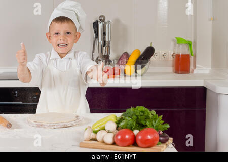 Begeisterter kleine Junge gekleidet in Köche Kleidung Kochen Pizza aufgeben ein Daumen des Erfolgs wie er die Basis für die Qualit beendet Stockfoto