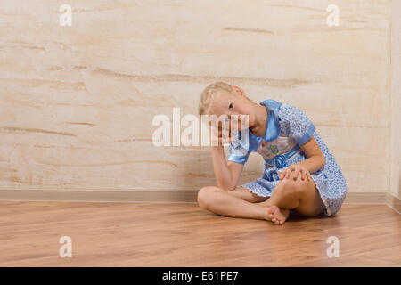 Gelangweilt kleines Mädchen barfuß sitzen mit gekreuzten Beinen auf einem Holzfußboden mit dem Kopf ruht auf ihrer Hand beobachten und warten mit Exemplar. Stockfoto