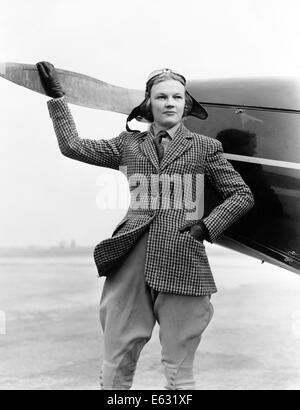 1930ER JAHREN FRAU AVIATOR PILOT STEHEN NEBEN FLUGZEUG EINERSEITS PROPELLER FLUG BRILLE CAP TWEED ANZUG MODE Stockfoto