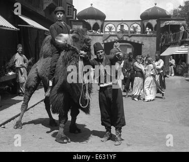 1910S 1920S VERLOR MAN IN BOWLER HUT REITEN BAKTRISCHEN KAMEL FRAGT RICHTUNGEN VON MENSCHEN IM ZENTRUM DER ARABISCHEN DORF STUMMFILM NOCH Stockfoto