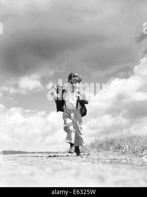 1940S 1950S JUNGE LÄUFT AUF KAMERA AUF LANDSTRAßE HOLDING STRAUß FRÜHLINGSBLUMEN IN EINER HAND Stockfoto