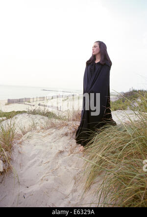 1960S 1970S TRAURIG DÜSTEREN EINSAME FRAU TRAGEN SCHWARZE LANGE VIKTORIANISCHEN CAPE STEHEN AM STRAND SAND DUNE Stockfoto