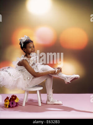 1970ER JAHREN AFROAMERIKANISCHE MÄDCHEN SITZEN AUF STUHL IN WEIßEN TUTU KOSTÜM BALLETTSCHUHE ANZIEHEN Stockfoto