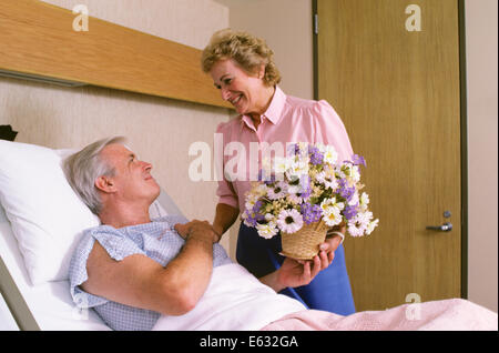 1980ER JAHRE FRAU GEBEN BLUMEN IM KRANKENHAUS MANN IM BETT LIEGEND Stockfoto