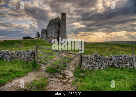 Magpie Mine, Derbyshire bei Sonnenuntergang, gesehen aus einem Feld mit Reflexionen in einer Pfütze unter dem Tor. Sonnenstrahlen scheinen durch die Wolken. Stockfoto