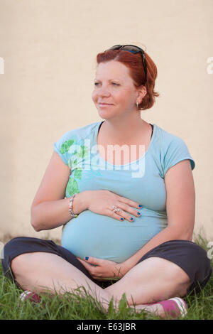 Glücklich Rothaarige schwangere Frau sitzen auf der Wiese, sonnigen Tag zu genießen. Stockfoto