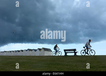 Gewitterwolken Brauen über Strandhütten und Boote an der Strandpromenade von Göring, West Sussex, wie Radfahrer und Menschen vorbeigehen Stockfoto