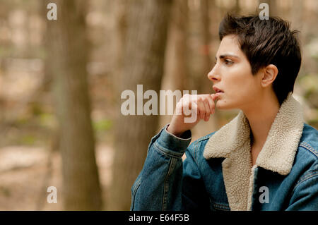 Eine schöne junge Frau mit kurzen, dunklen Haaren und trägt eine Sherpa-gefütterte Jeansjacke stellt nachdenklich in den Wäldern. Stockfoto
