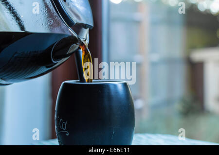 Heißer dunkler Filterkaffee, der aus einer Glaskaraffe in eine schwarze Kaffeetasse oder einen Becher gegossen wird. Terrassentüren, die zu einem Garten im Hintergrund führen. Stockfoto