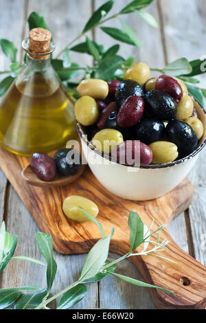 Marinierte Oliven (grün, schwarz und lila) in Keramikschale und Holzlöffel mit Flasche Olivenöl gemischt. Selektiven Fokus. Stockfoto