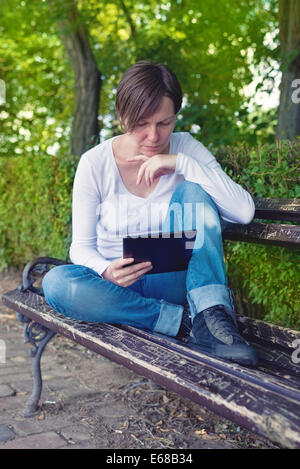 Frau liest E-Book auf digitale Tablettcomputer sitzend auf einer Holzbank im Park. Stockfoto