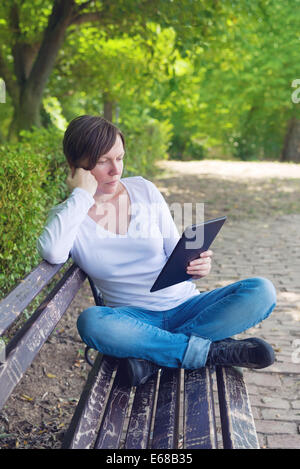 Frau liest E-Book auf digitale Tablettcomputer sitzend auf einer Holzbank im Park. Stockfoto